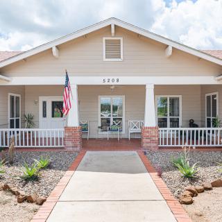 A charming single-story house with a front porch and American flag, surrounded by light landscaping.