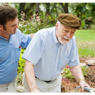 A caregiver assists an elderly man walking in a garden.