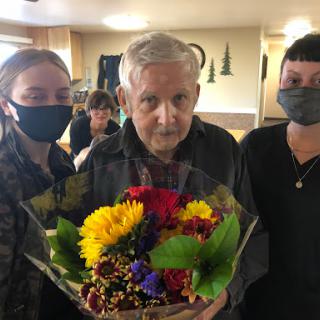 An elderly man surrounded by two individuals, holding a vibrant bouquet of flowers in a warm indoor setting.
