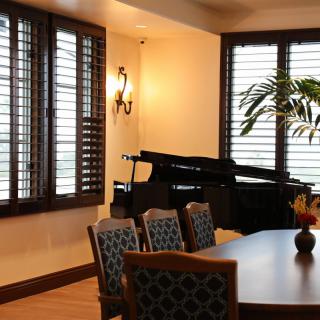 A cozy dining room with a grand piano and elegant furnishings near large windows with shutters.