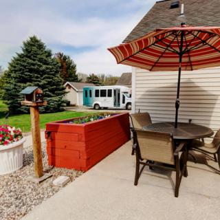 A cozy patio with an umbrella and seating, overlooking a suburban garden with a bus parked in the background.