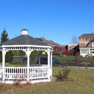 A white gazebo in a grassy area with buildings in the background under a clear blue sky.