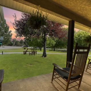 A tranquil evening view from a porch with rocking chairs, overlooking a lush lawn and trees at sunset.