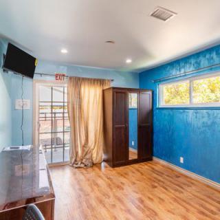 A bright room with blue walls, a wooden floor, and simple furnishings including a wardrobe, desk, and mounted TV.
