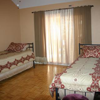 A cozy bedroom featuring two single beds with patterned bedding, illuminated by natural light streaming through large curtains on a wooden floor.