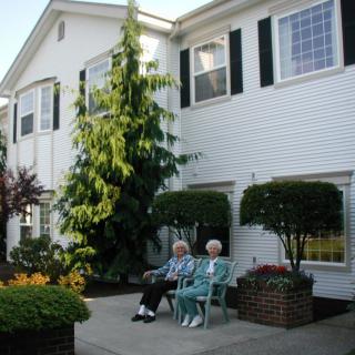 Two elderly individuals sitting on chairs outside a residential building, surrounded by greenery.