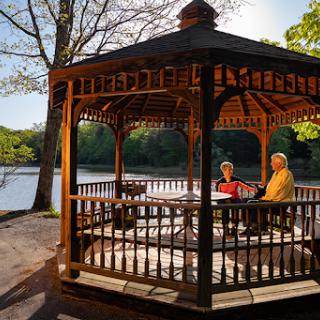 A serene afternoon in a gazebo by the lake, where two individuals enjoy a peaceful moment surrounded by nature.