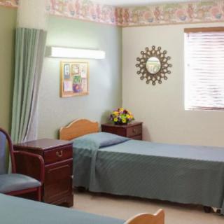 A hospital room with two neatly made beds, a small table with a flower arrangement, and a window allowing natural light to enter.