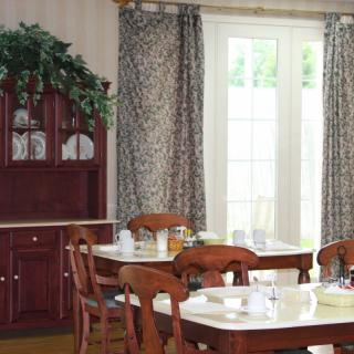 A cozy dining room featuring a wooden table set with dishes and surrounded by chairs, with a cabinet in the background and patterned curtains framing the windows.