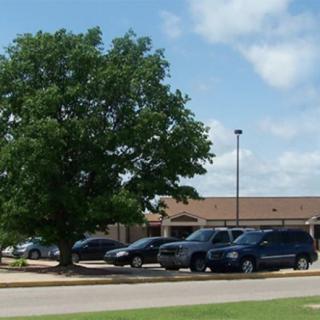 A parking lot with vehicles in front of a building, alongside a large tree under a partly cloudy sky.