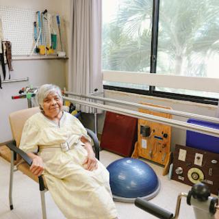 An elderly individual seated in a rehabilitation center, surrounded by various exercise equipment.