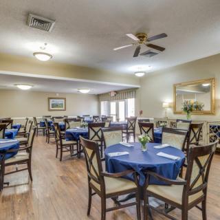 A spacious dining room with neatly arranged tables and chairs, featuring blue tablecloths and a ceiling fan.