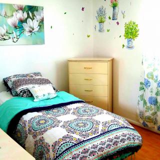 A cozy bedroom with a single bed, featuring colorful and floral bedding, complemented by wall art and a wooden dresser under natural light.