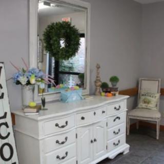 A welcoming entryway with a white dresser, decorative mirror, wreath, and a welcome sign.