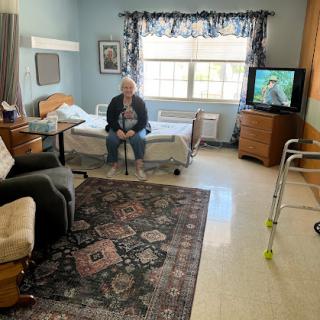 An elderly person sitting in a well-furnished hospital room with a view outside.