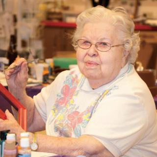 An elderly woman engaged in painting, surrounded by art supplies.