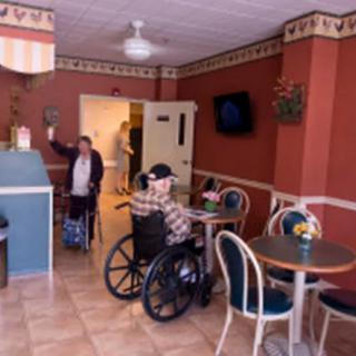 Two senior citizens are socializing inside a cafe, one seated at a table and the other standing with a walker.