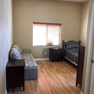 A small bedroom with a wooden floor, featuring a bed, couch, nightstand, and window.