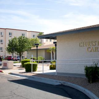 A view of a residential building with a parking area and green landscaping under a clear blue sky.