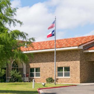 A brick building with American and Texas flags in front, surrounded by trees and under a cloudy sky.