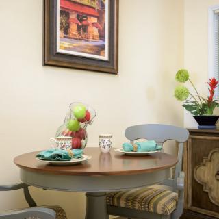 A cozy dining nook with a round table set for two, featuring a decorative fruit bowl and framed artwork on the wall.