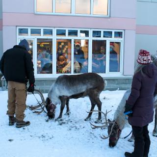 Two reindeer being handled by a couple of people in snowy conditions outside a building.