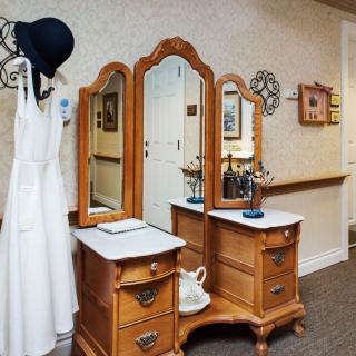 A vintage wooden dresser with a triple mirror, accompanied by a hanging hat and dress in an elegant interior setting.