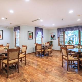 A well-lit dining room with tables and chairs, featuring hardwood floors and windows with curtains.