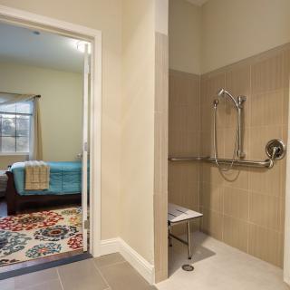 An accessible bathroom with a walk-in shower featuring grab bars and a shower chair, adjacent to a cozy bedroom with bright decor.