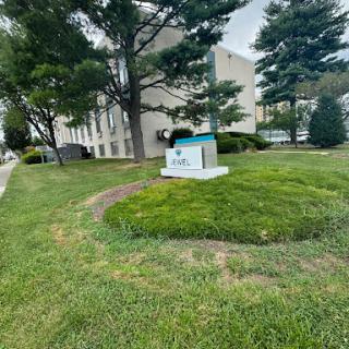 A view of a landscaped area with a sign in front of a building, surrounded by trees and green grass.