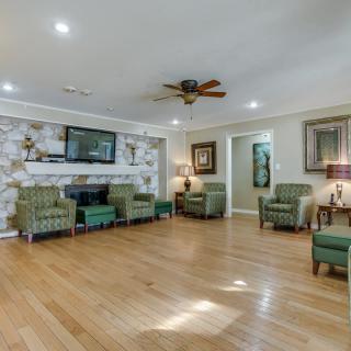 A spacious living room with wooden flooring, green armchairs, a ceiling fan, and a television mounted above a decorative fireplace.