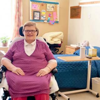 An elderly woman smiling while sitting in a wheelchair in a nursing home bedroom.