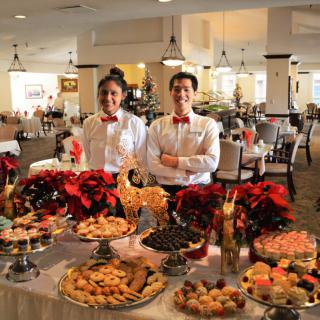Two waiters stand behind a festive holiday dessert buffet in a decorated restaurant.