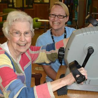 A joyful senior woman participates in a fitness session, supported by a staff member.