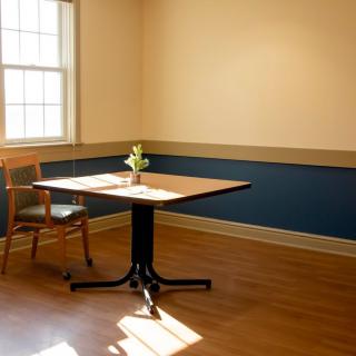 A minimalist room with a single table and chair near a window, accented by natural light and a small flower vase.