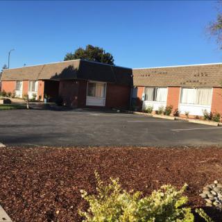 A single-story brick building with a parking lot in front, surrounded by landscaping under a clear blue sky.