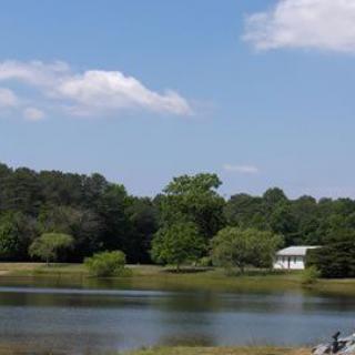 A serene view of a lake surrounded by trees, with a small house in the distance under a partly cloudy sky.