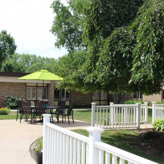 A serene outdoor patio area with a table, chairs, and a green umbrella, surrounded by trees and a white fence.