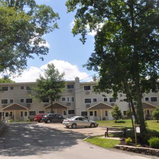 A residential building with a parking lot and cars on a sunny day, surrounded by trees.