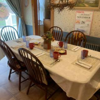 A dining table set for a meal with six chairs, featuring a white tablecloth, red mugs, and neatly placed napkins.