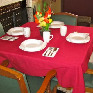 A neatly set dining table with a red tablecloth, featuring plates, mugs, cutlery, and a floral centerpiece.