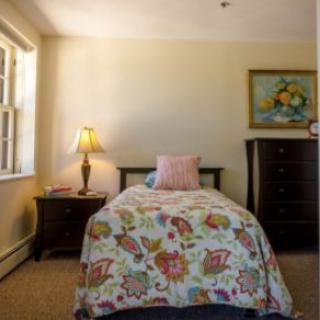 A cozy bedroom with a single bed featuring floral bedding, flanked by a lamp on a nightstand and a dresser with artwork above.