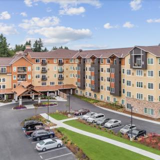 A multi-story residential building with a parking lot and surrounding greenery under a partly cloudy sky.