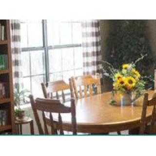 A cozy dining area with a wooden table and chairs, adorned with a vase of sunflowers. Natural light streams in through the window, and a bookshelf lines the wall.