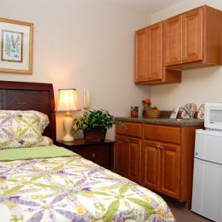 A cozy bedroom with a single bed, lamp, and kitchen cabinets.