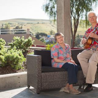 An elderly couple enjoying a sunny day on the patio, with one playing guitar.
