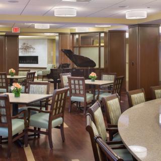 A well-appointed dining room featuring tables and chairs, with a piano in the background and floral decorations on the tables.