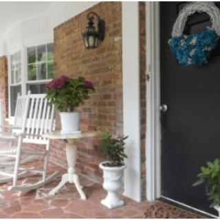A cozy porch featuring a white rocking chair, potted plants, and a decorative wreath on the front door.