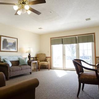 A cozy living room with natural light filtering through large windows, featuring comfortable seating and a ceiling fan.