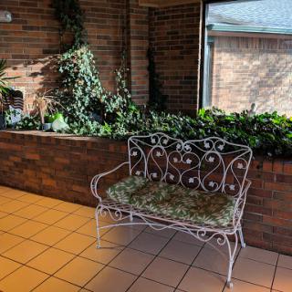 A decorative metal bench surrounded by lush indoor plants and a brick wall, illuminated by natural light.